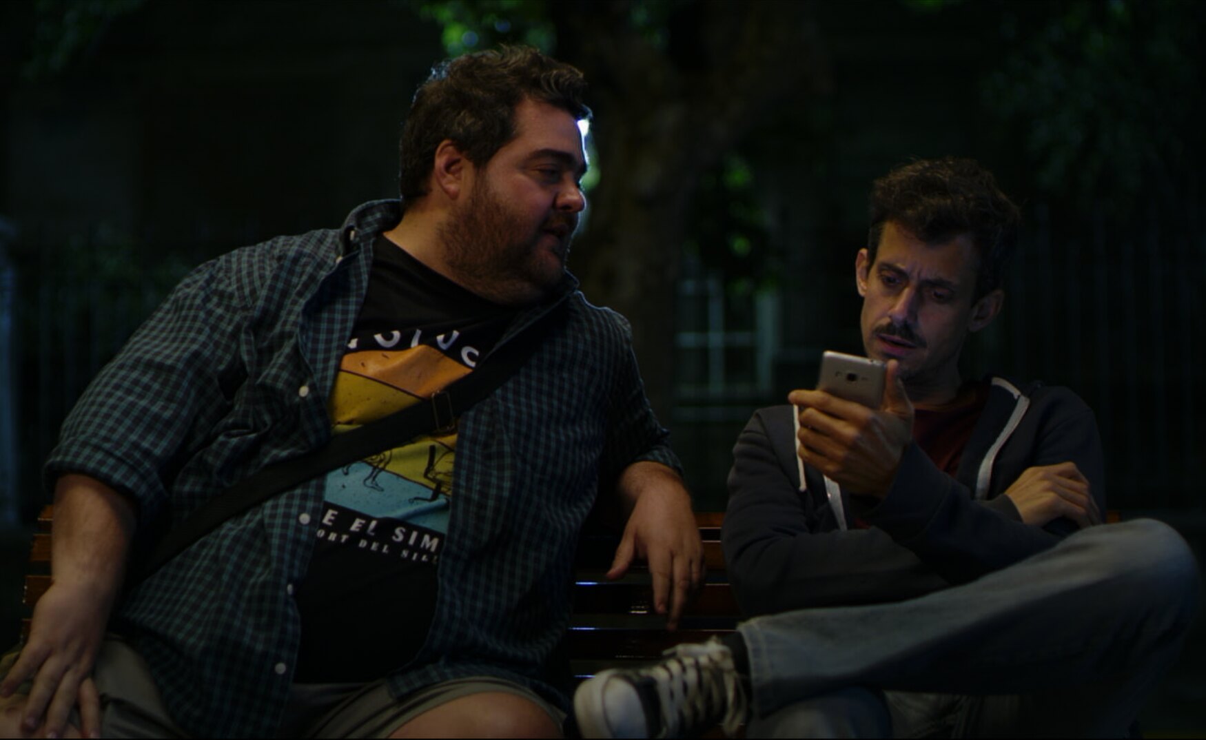 Hernán y Chiri sentados en una plaza, de noche, mirando un teléfono
